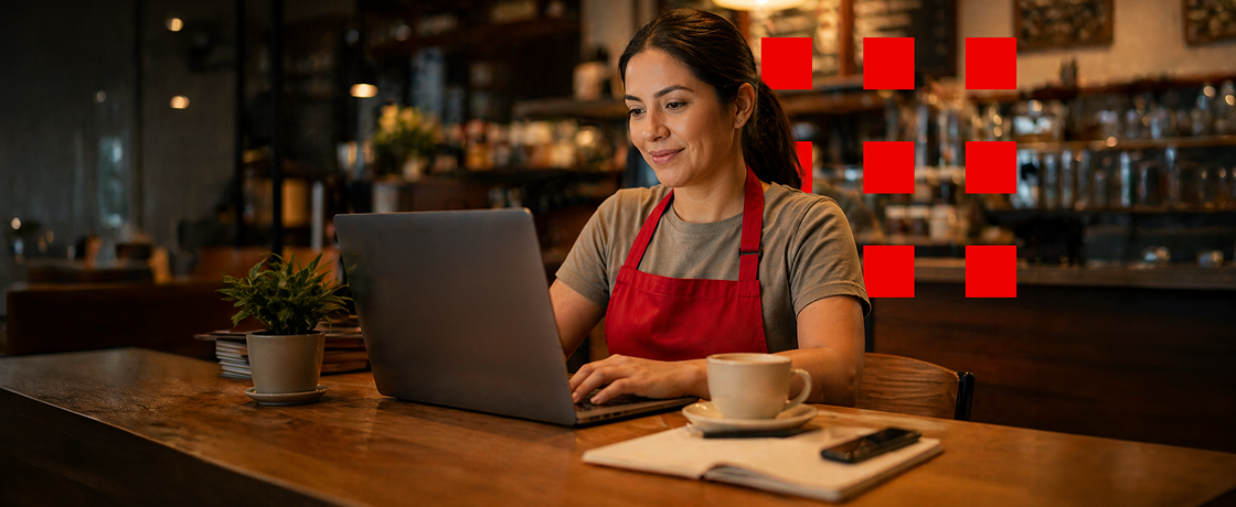 Una mujer con un delantal rojo trabajando con el laptop en una cafetería, a la derecha aparece una taza de café